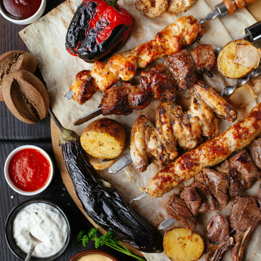 Grillplatte mit Spießen, Gemüse und Saucen auf dunklem Holztisch